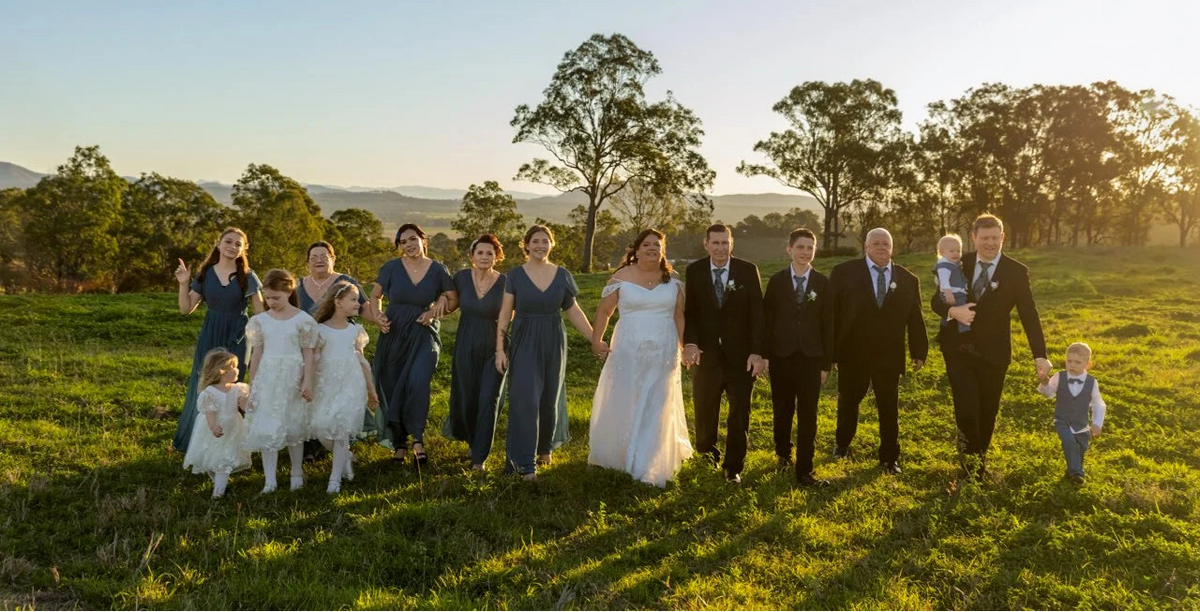 A wedding party of 12 people posing together outdoors in a grassy field at sunset. The bride wears a white wedding gown, the groom wears a black suit, and the bridesmaids wear slate blue dresses, while the groomsmen wear black suits.