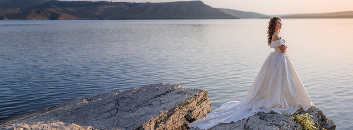 Woman in a white off-shoulder wedding dress standing on a rocky outcrop with a scenic background.