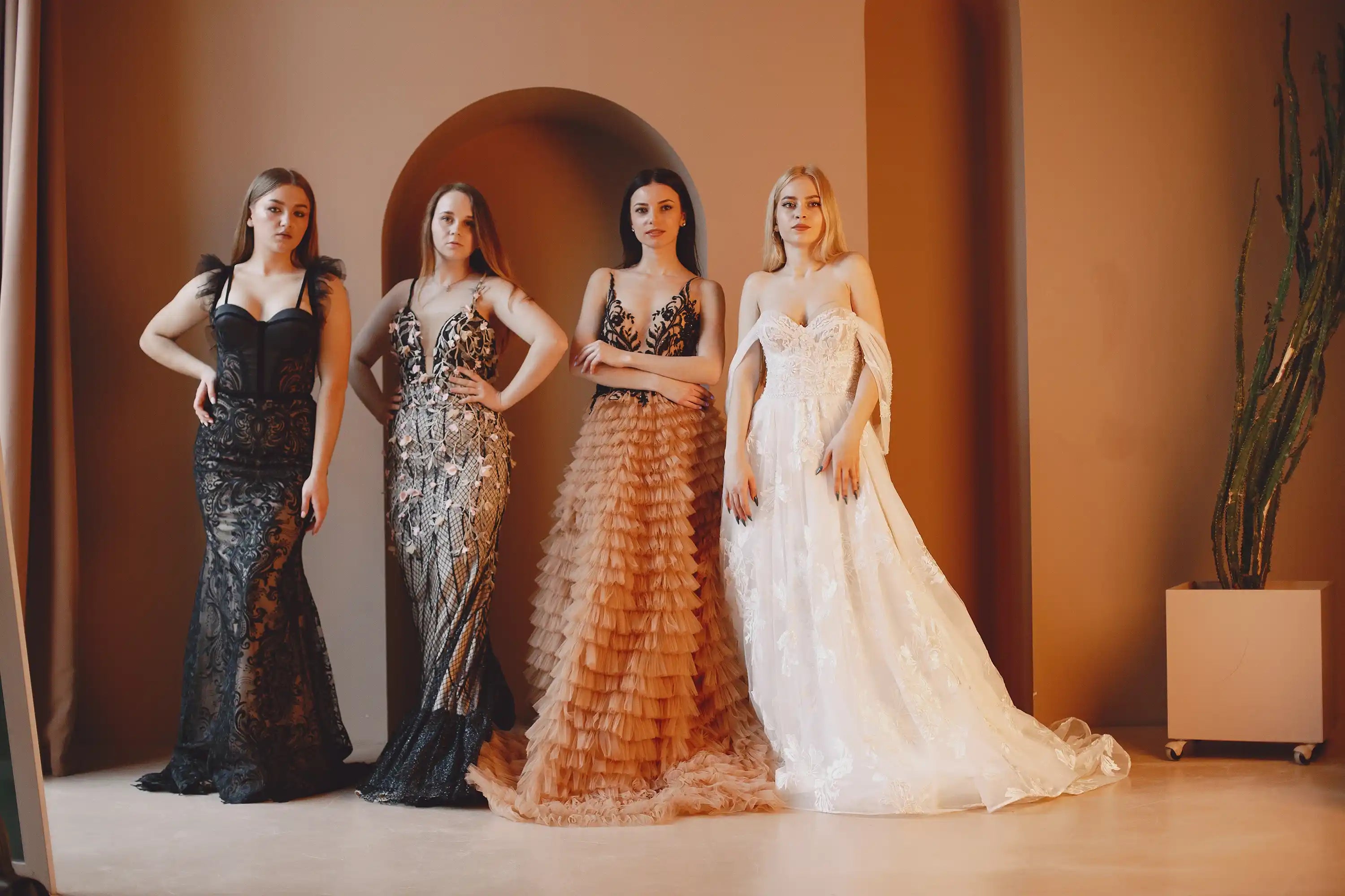 Four women wearing elegant formal evening gowns including black lace mermaid dress, gold sequin gown, champagne tiered ruffle formal dress and white lace ball gown